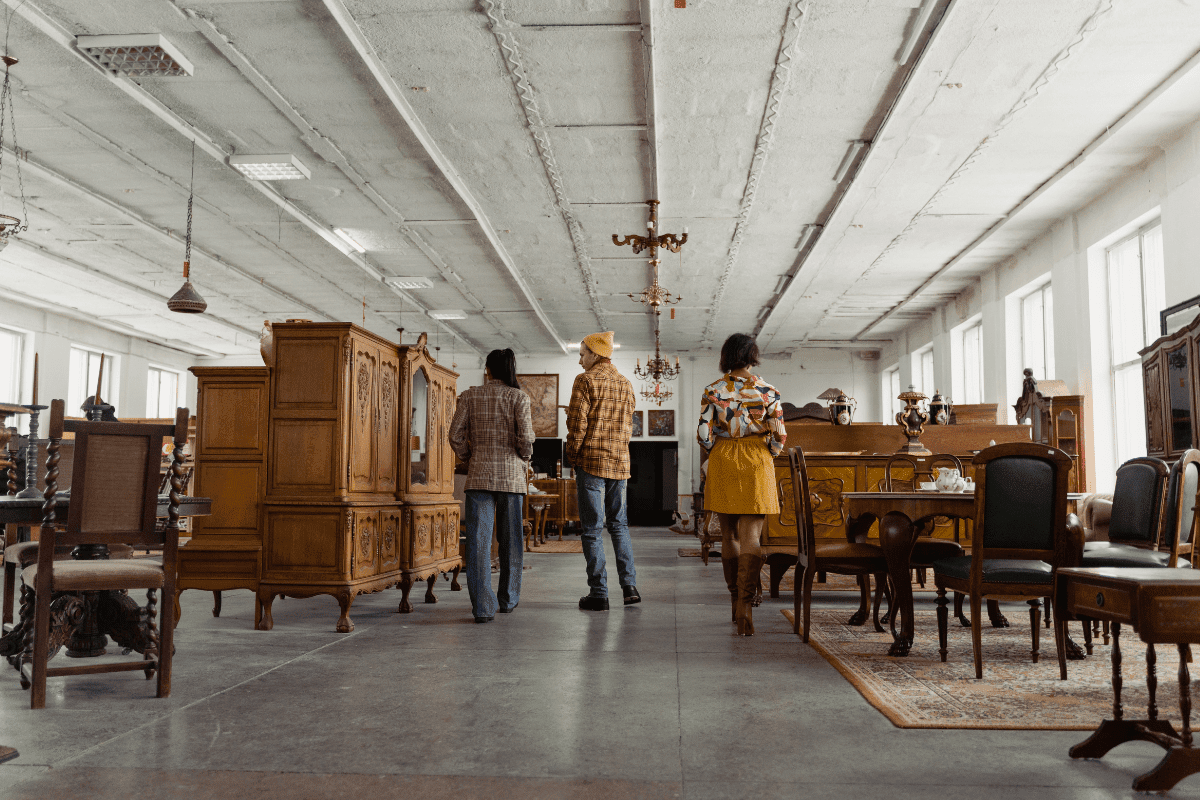 Three people stand in a spacious, well-lit antique furniture store, surrounded by wooden cabinets, tables, and chairs, as they examine the items on display.