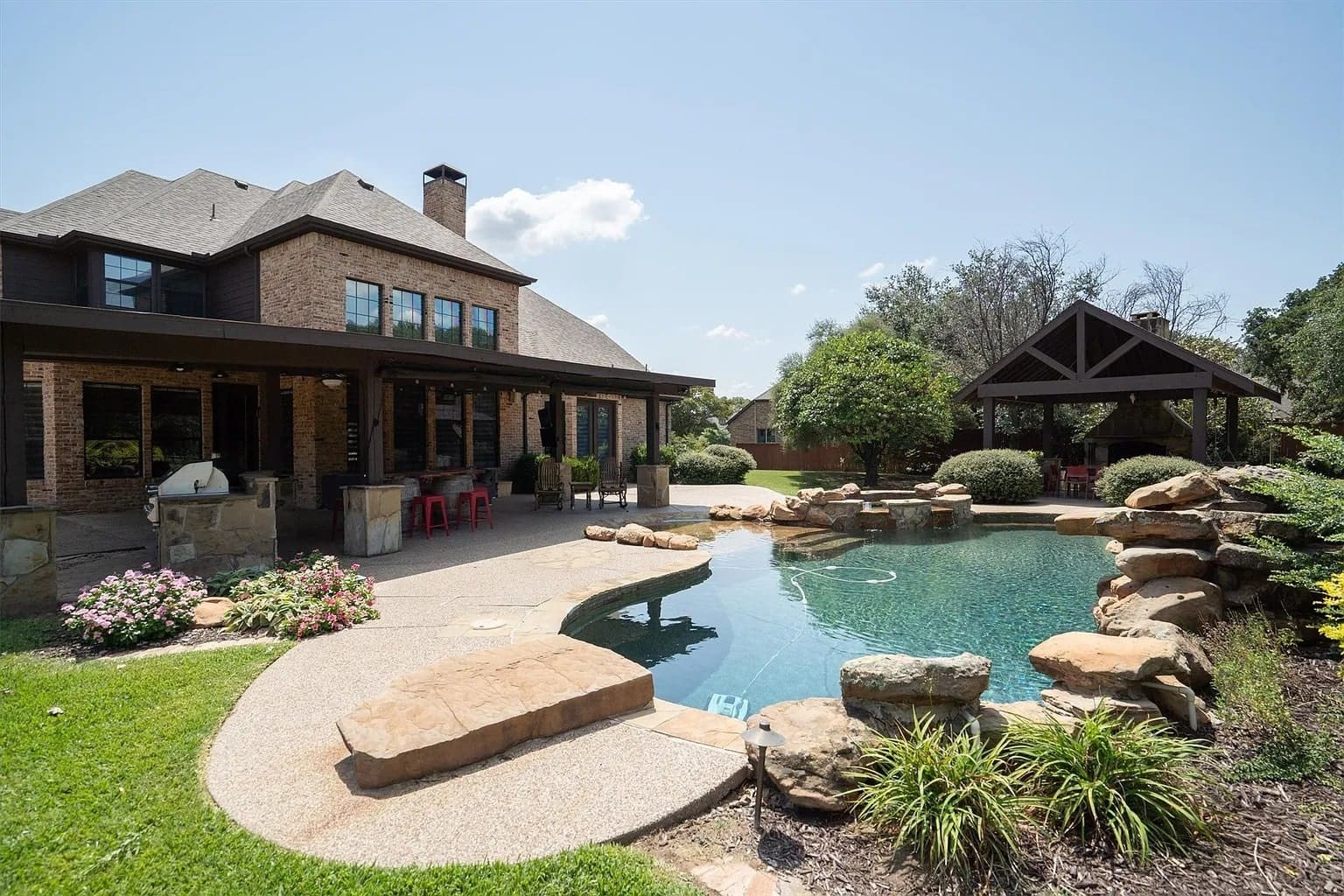Backyard with a pool, patio, and gazebo, surrounded by greenery and a large house.
