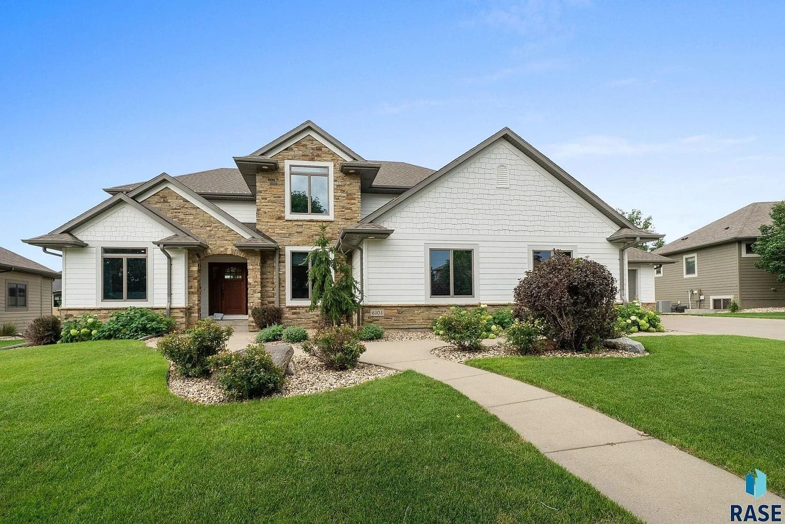 Modern suburban house with stone and white siding, surrounded by a well-maintained lawn and shrubs.
