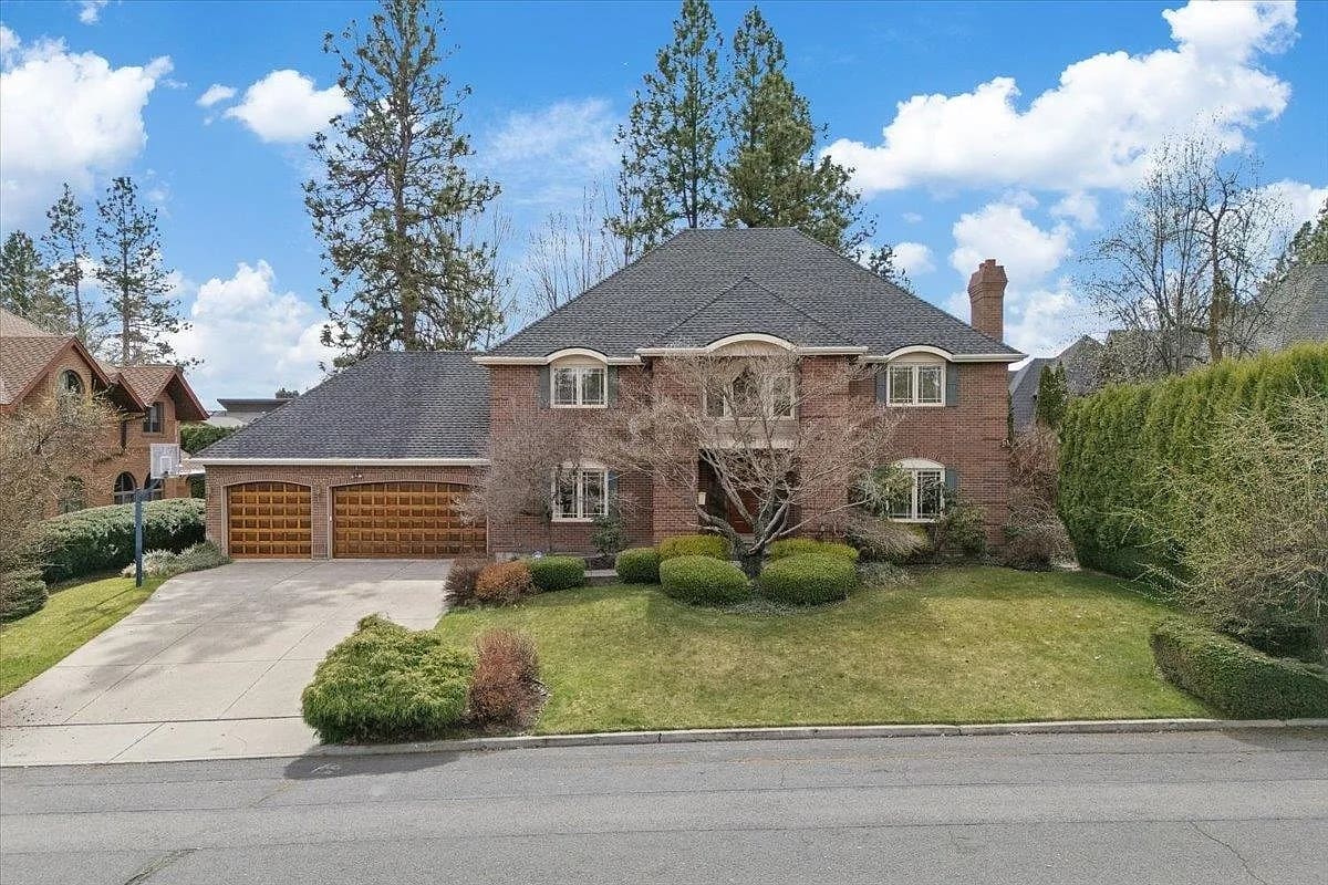 Large brick house with a three-car garage, surrounded by trees and a well-maintained lawn.
