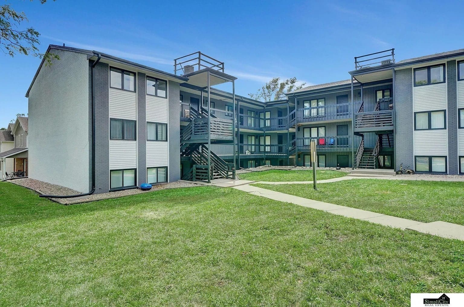 Three-story apartment building with balconies, surrounded by grass and walkways under a clear blue sky.
