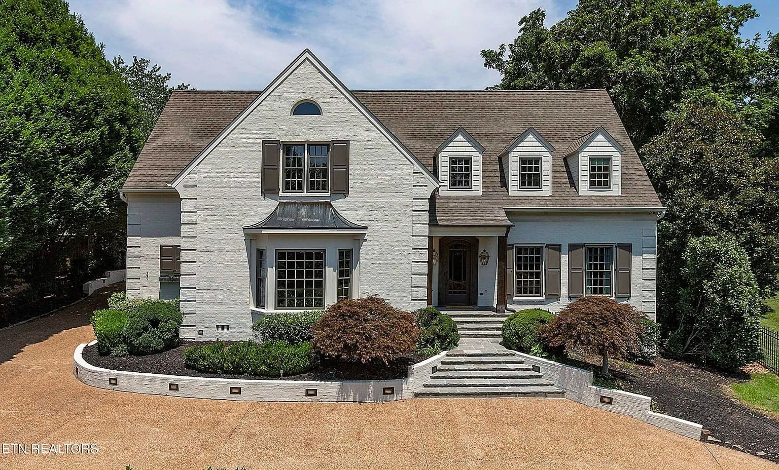 Two-story white brick house with dormer windows, surrounded by trees and landscaped shrubs.