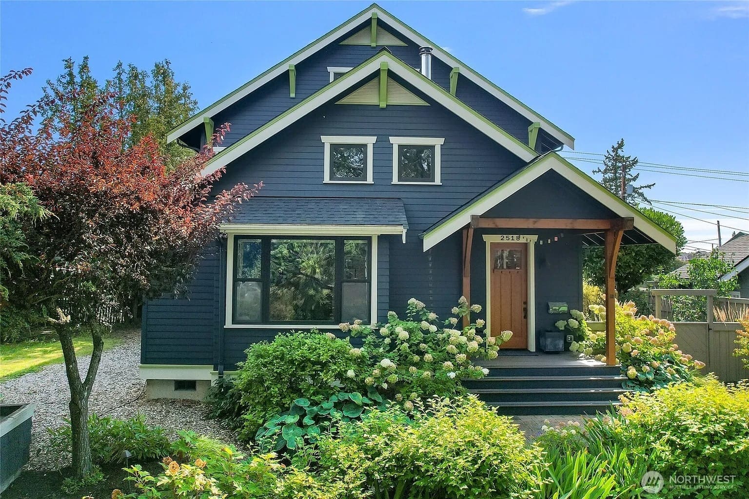 Blue two-story house with a wooden door, surrounded by greenery and a small garden.
