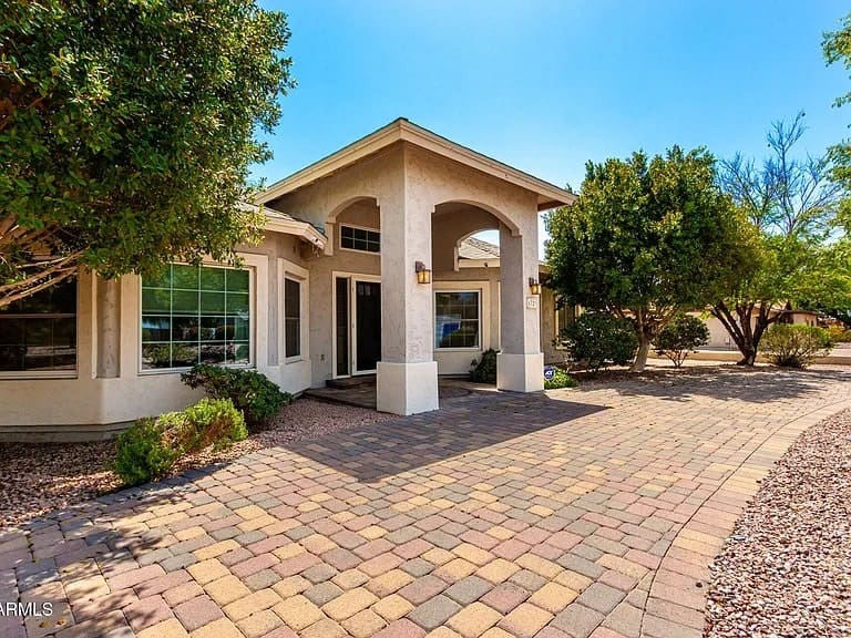 Single-story house with arched entrance, surrounded by trees, and a paved driveway.
