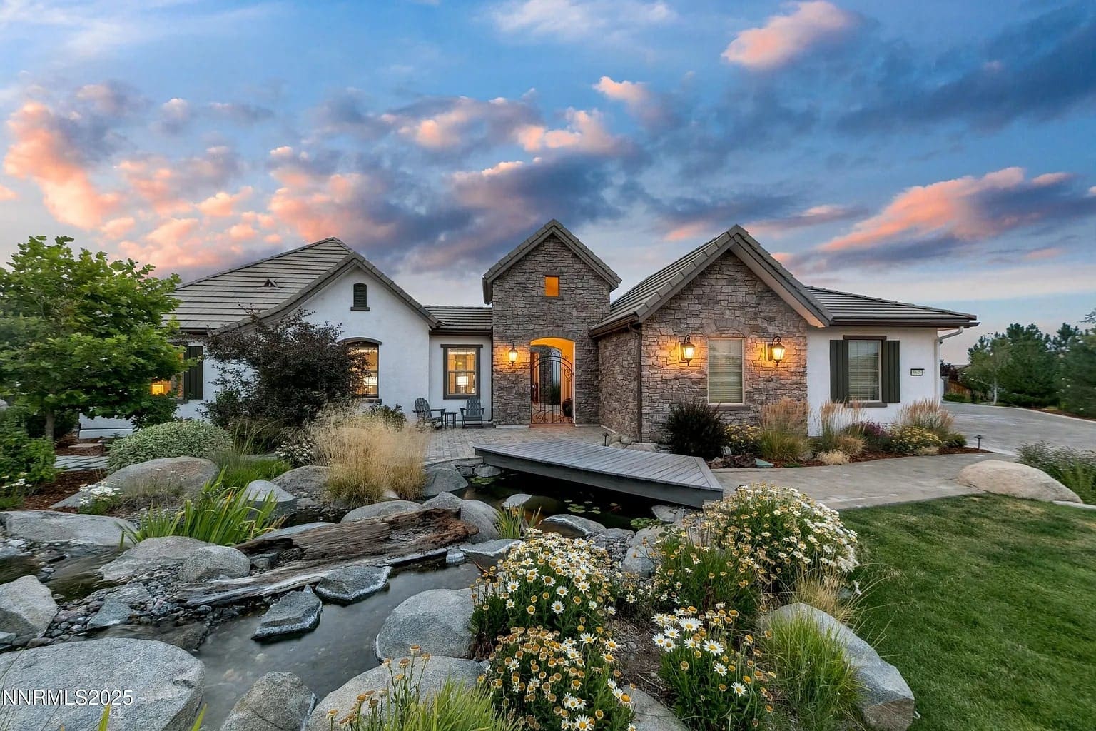 Single-story house with stone facade, lit exterior, garden, and small pond at sunset.