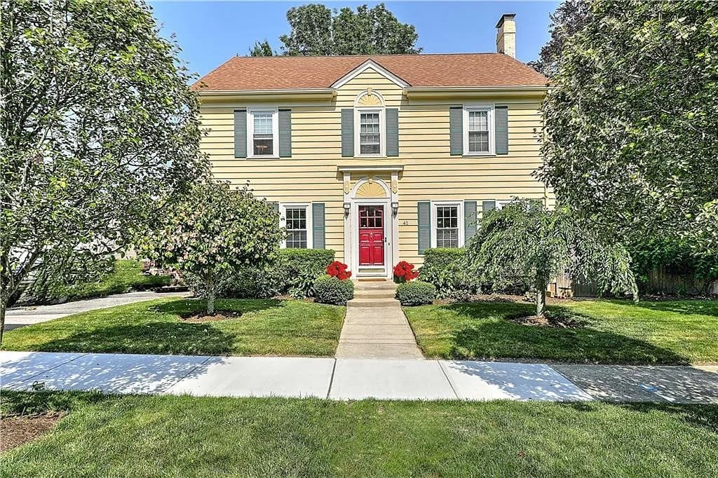 Yellow two-story house with red door, surrounded by trees and a well-kept lawn.
