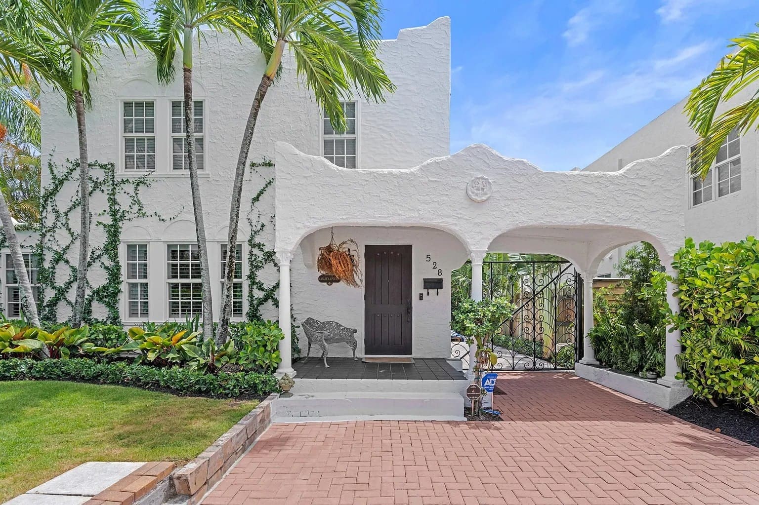 White stucco house with arched entryway, surrounded by palm trees and greenery.
