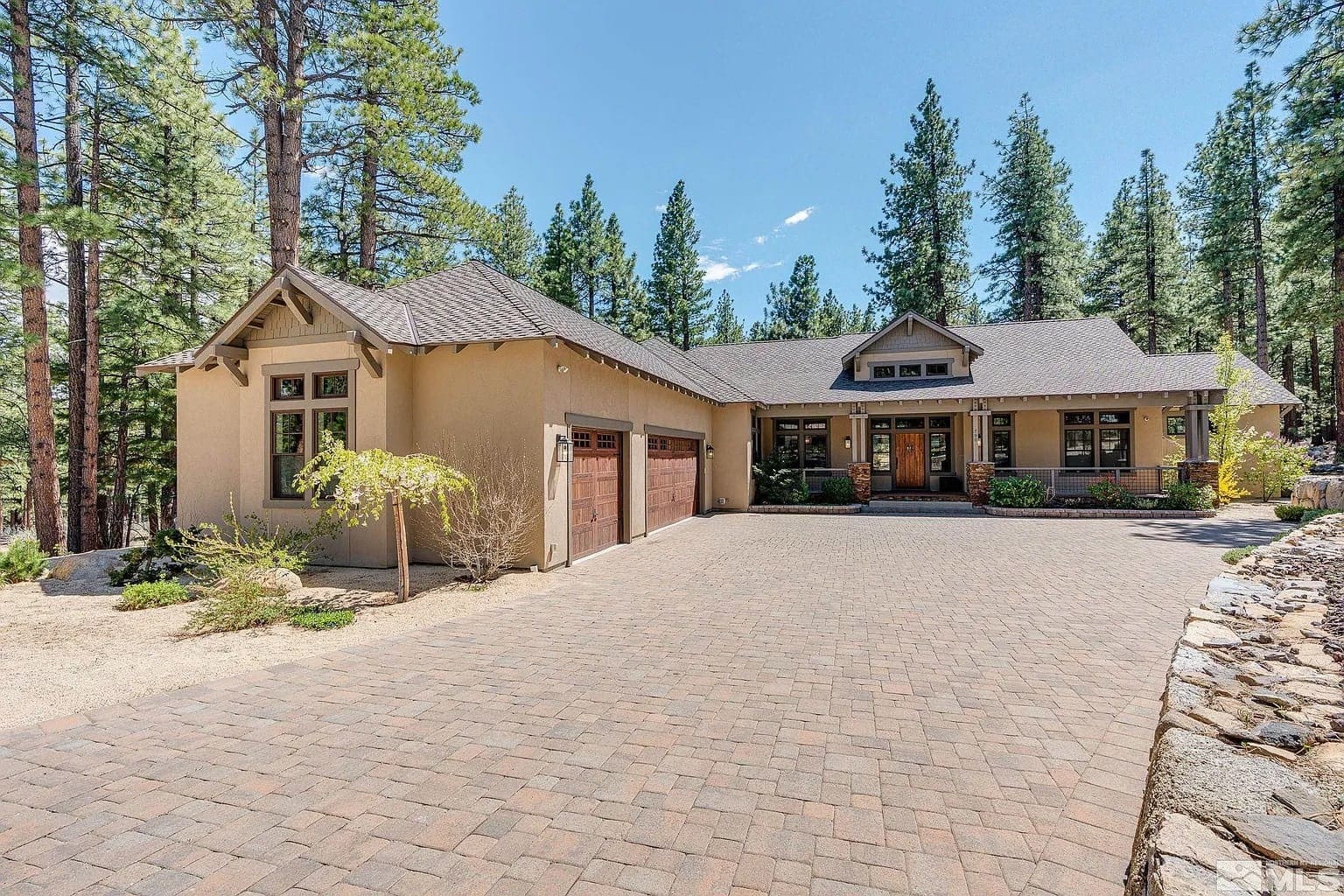 Single-story house with a three-car garage, surrounded by tall pine trees and a paved driveway.