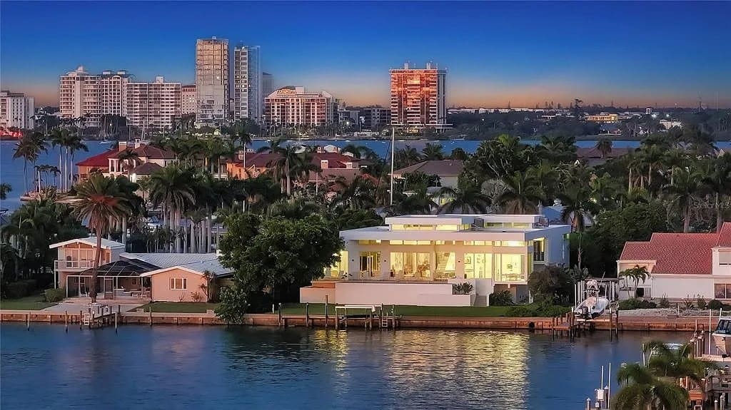 Modern waterfront house with large windows, surrounded by palm trees, city skyline in the background.