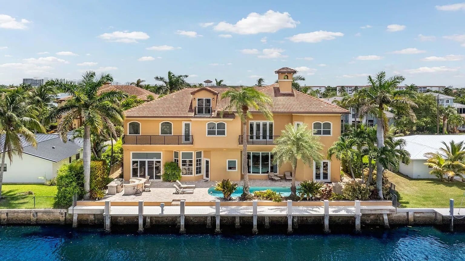 Large waterfront house with palm trees, pool, and patio, set against a clear blue sky.
