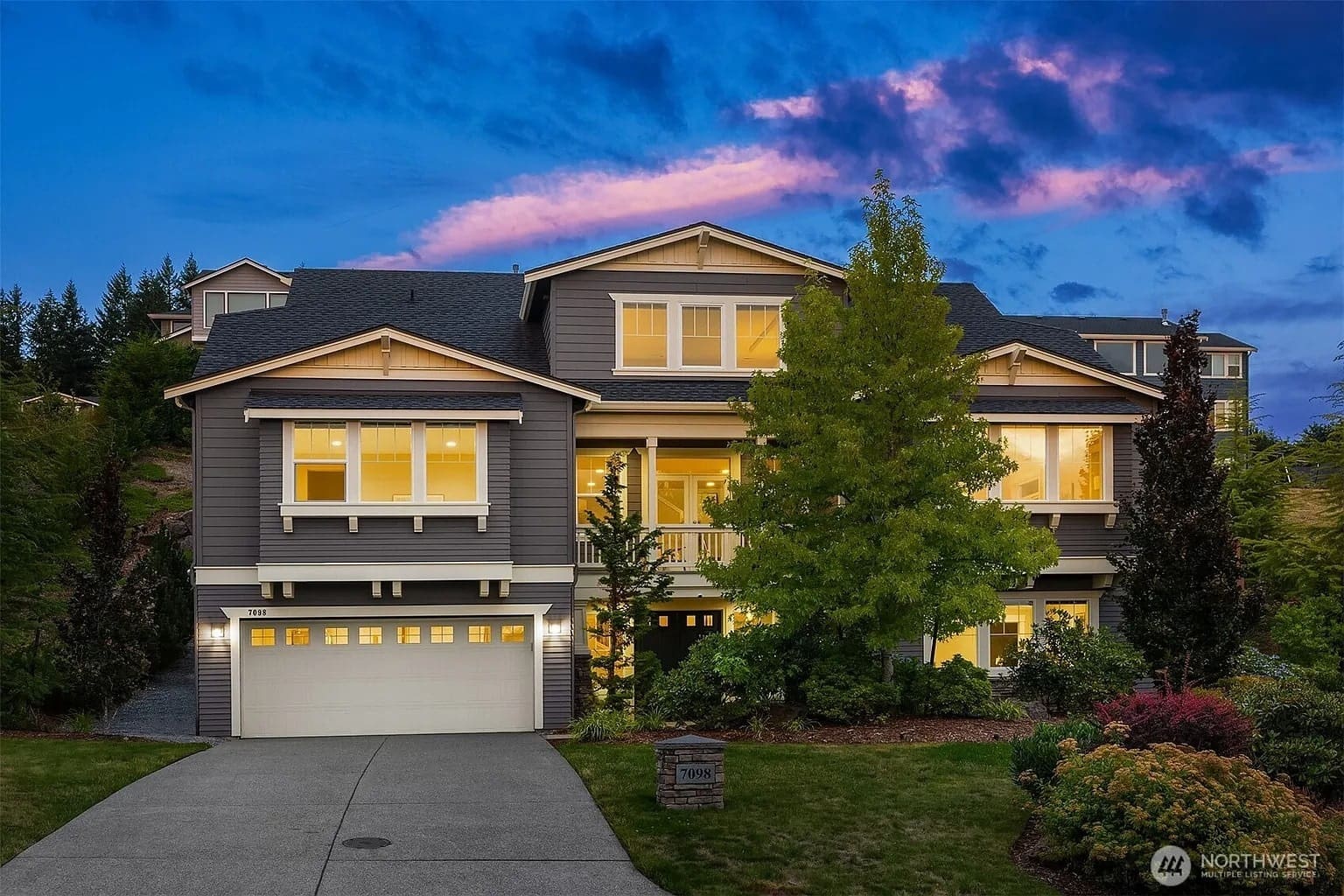 Large, illuminated two-story house with a landscaped yard, set against a vibrant sunset sky.
