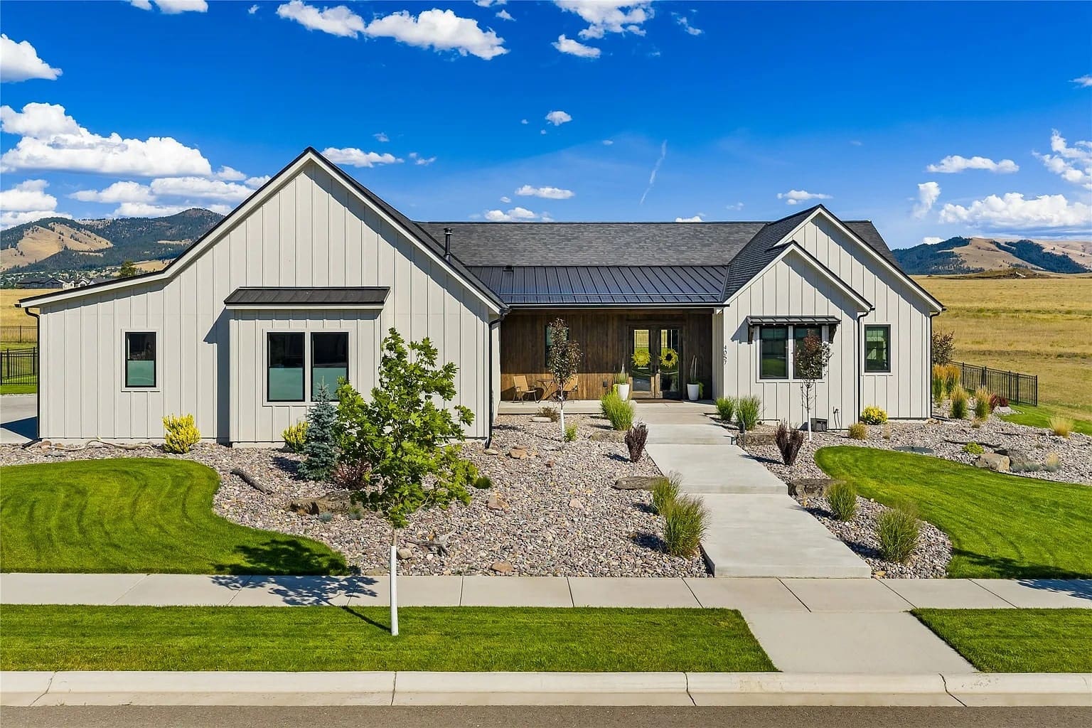 Modern single-story house with gray siding, landscaped yard, and mountain backdrop under a blue sky.
