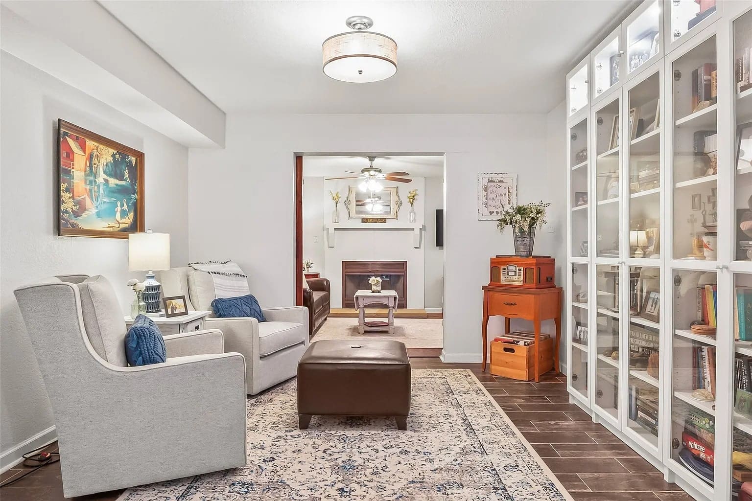 Cozy living room with armchairs, a brown ottoman, glass cabinet, and a fireplace in the background.
