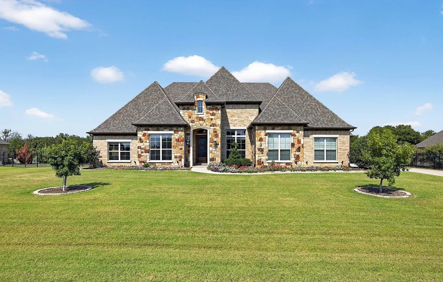 Large stone house with multiple peaks, surrounded by a well-maintained lawn and small trees.

