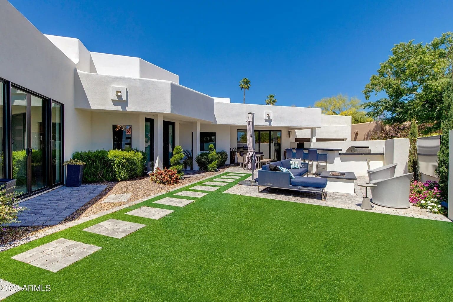 Modern backyard with artificial grass, patio furniture, and a built-in grill against a white house.