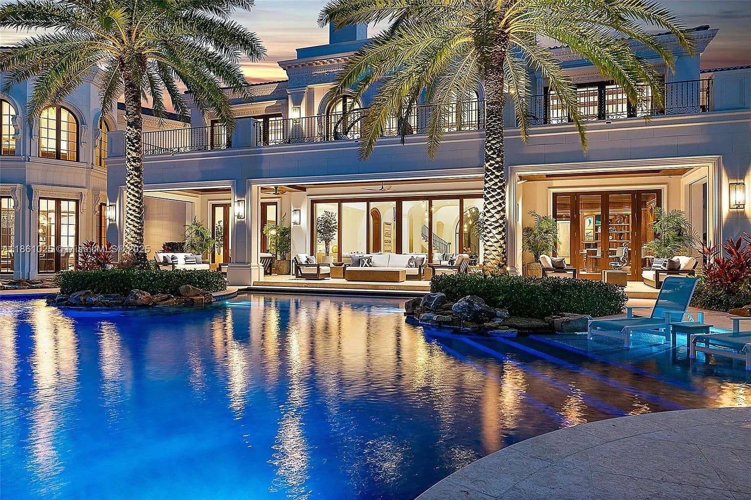 Luxurious mansion with large pool, palm trees, and elegant outdoor seating at sunset.
