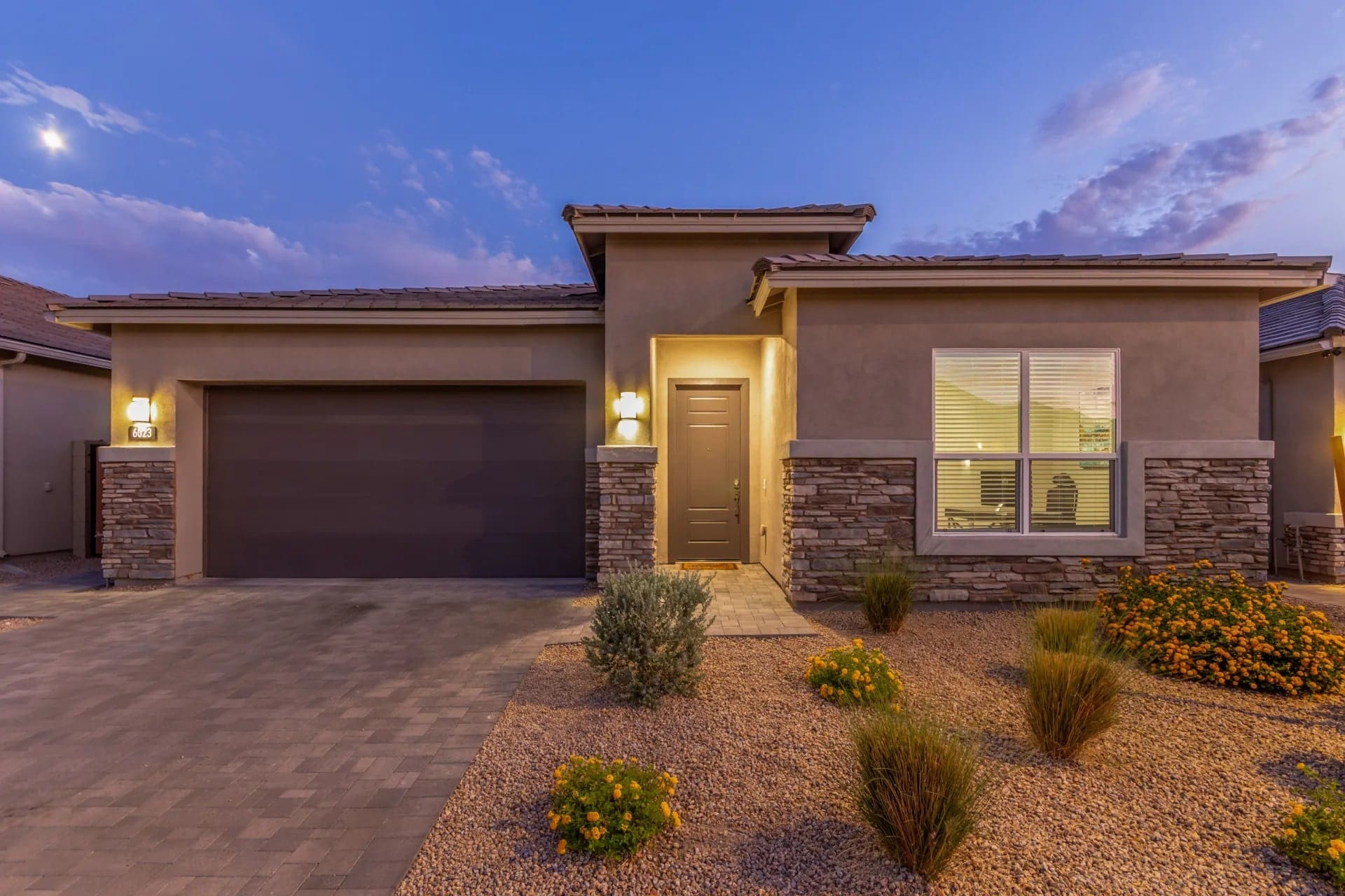 Modern single-story house with stone accents, two-car garage, and desert landscaping at dusk.