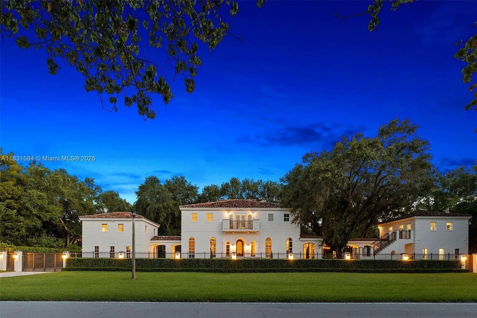 Large, elegant mansion with bright exterior lights, surrounded by trees, under a deep blue evening sky.