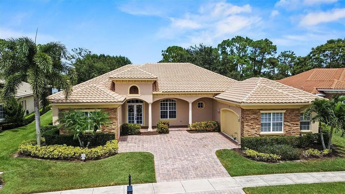 Single-story house with a tiled roof, brick driveway, and surrounding greenery under a blue sky.
