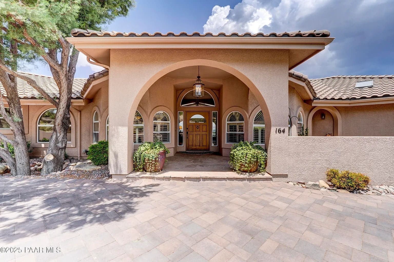 Single-story house with arched entrance, wooden door, and tiled roof, surrounded by trees and paved driveway.
