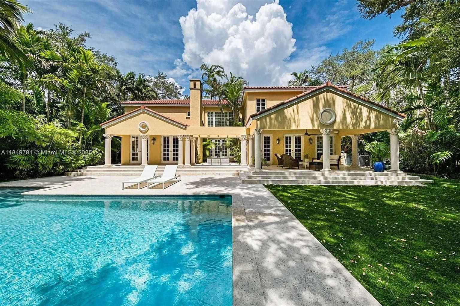 Yellow house with red roof, large patio, pool, and surrounded by lush greenery.