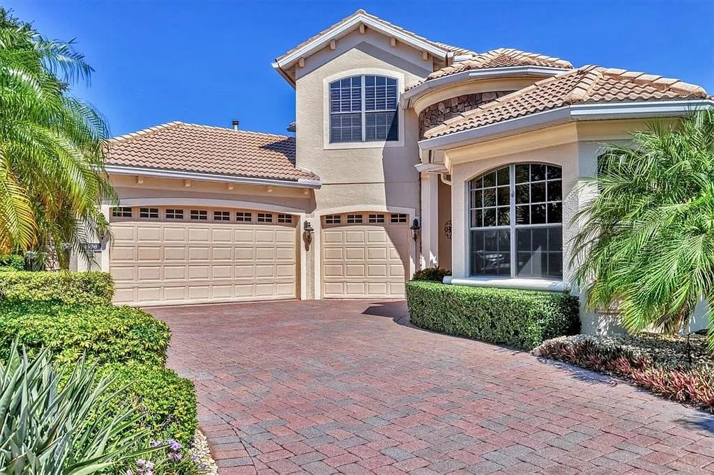 Large beige house with two garages, tiled roof, and landscaped driveway surrounded by greenery.
