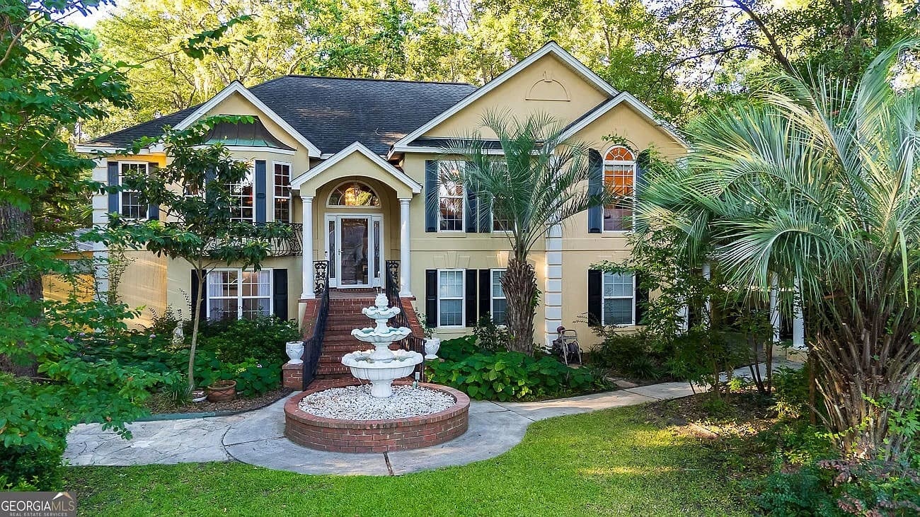 Two-story yellow house with a central staircase, surrounded by trees and featuring a white fountain.
