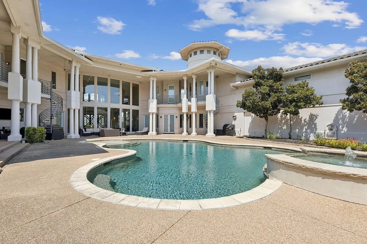 Luxurious mansion with a large circular swimming pool, surrounded by columns and landscaped gardens.
