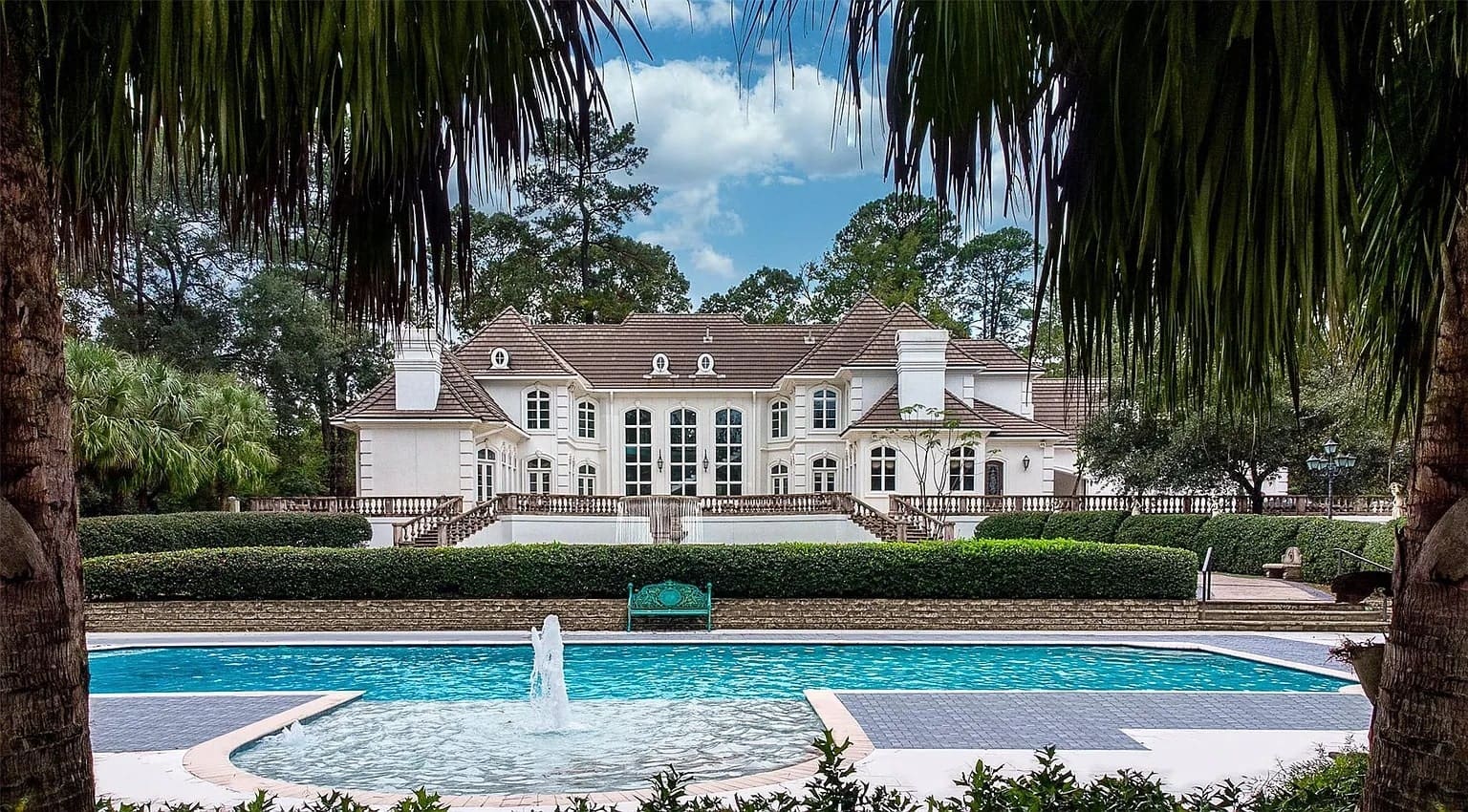 Large mansion with multiple chimneys, viewed from behind a pool with a central fountain.
