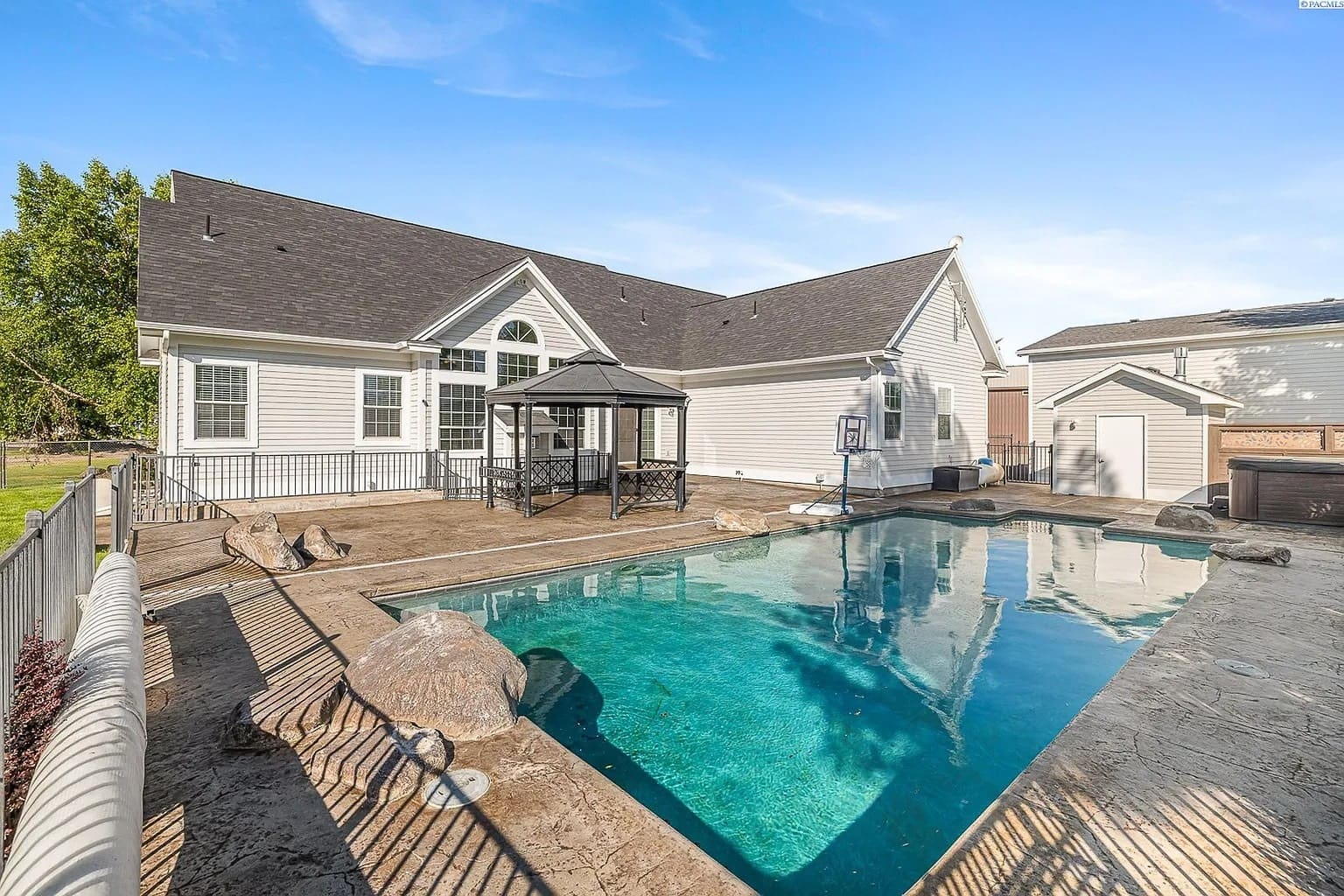 Backyard with a swimming pool, gazebo, and a white house under a clear blue sky.
