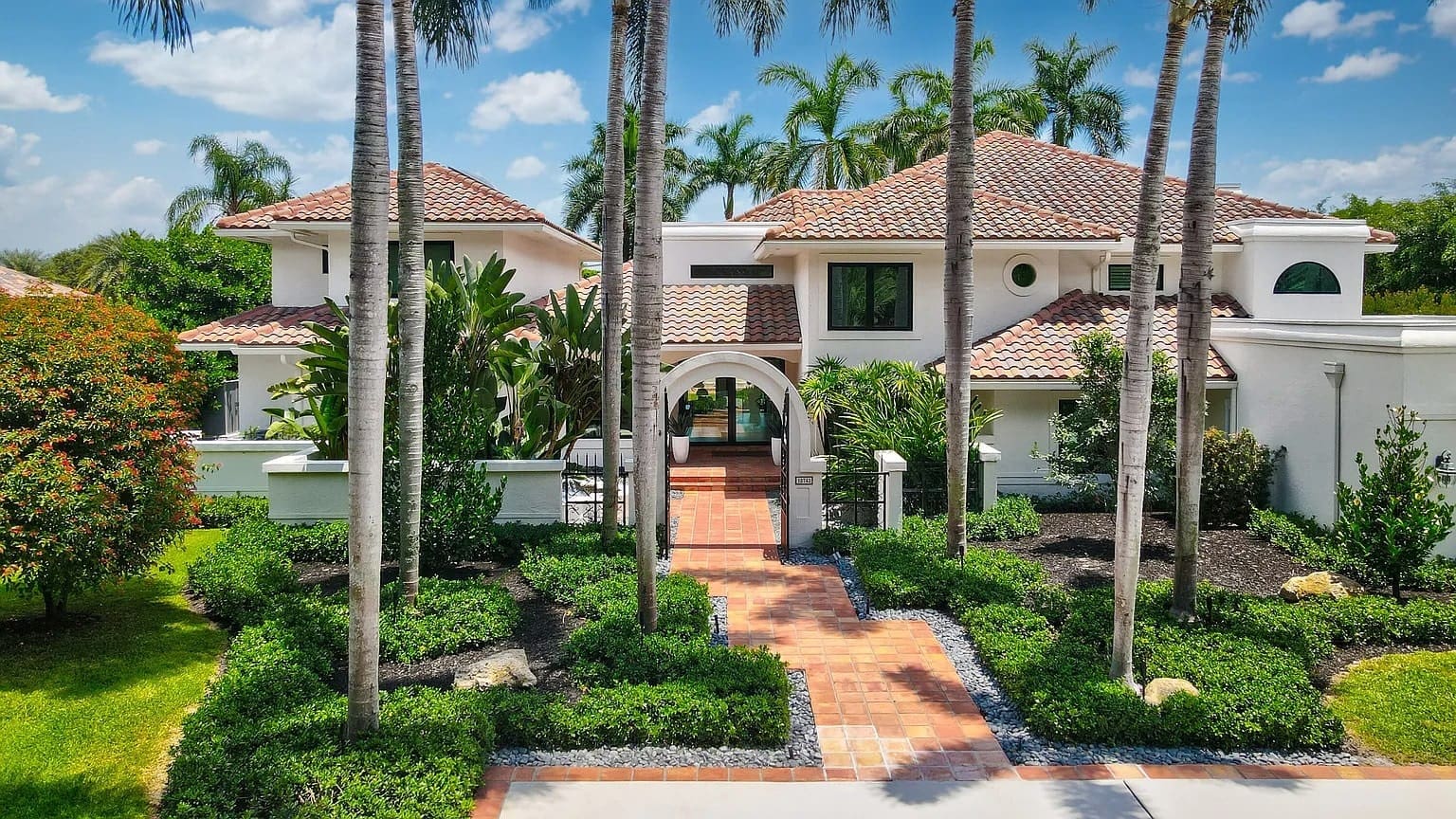 Mediterranean-style house with red-tiled roof, palm trees, and lush landscaping under a blue sky.