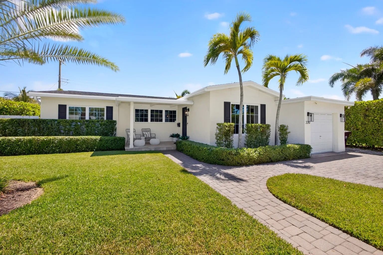 Single-story white house with palm trees, manicured lawn, and paved driveway under a clear blue sky.
