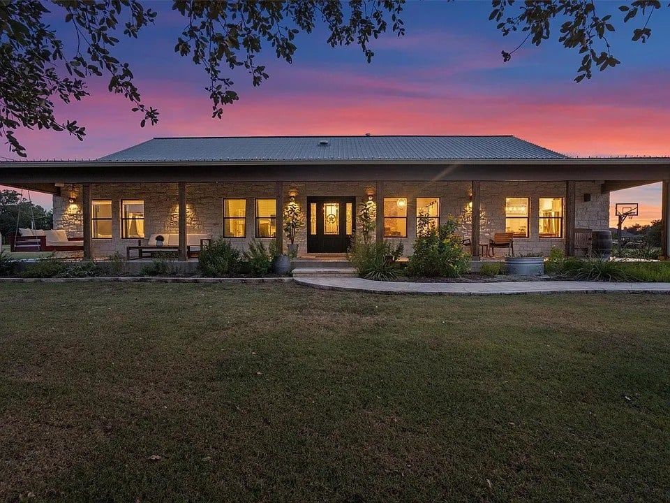 Single-story house with illuminated windows at sunset, surrounded by a grassy yard and trees.