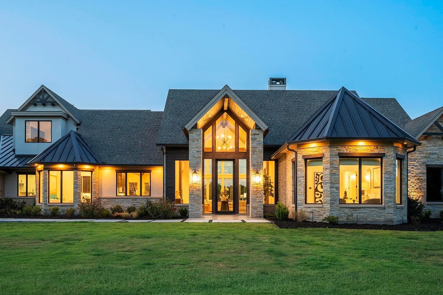 Modern house with large windows, stone exterior, and illuminated interior, set against a twilight sky.