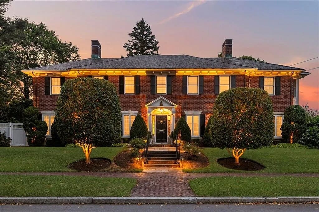 Two-story brick house with lit windows and manicured trees, set against a sunset sky.