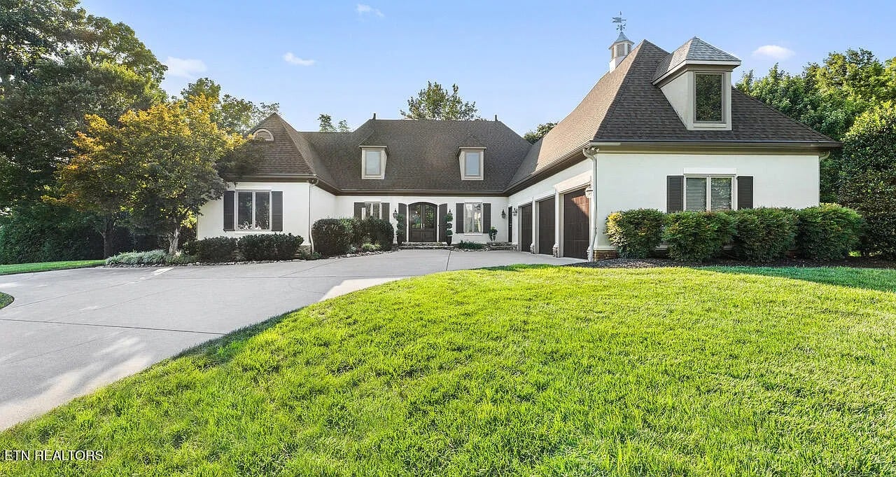Large suburban house with a sloped roof, circular driveway, and surrounding greenery.