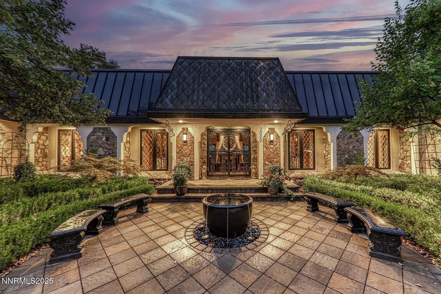 Elegant stone house with a central fountain, surrounded by benches and greenery at sunset.