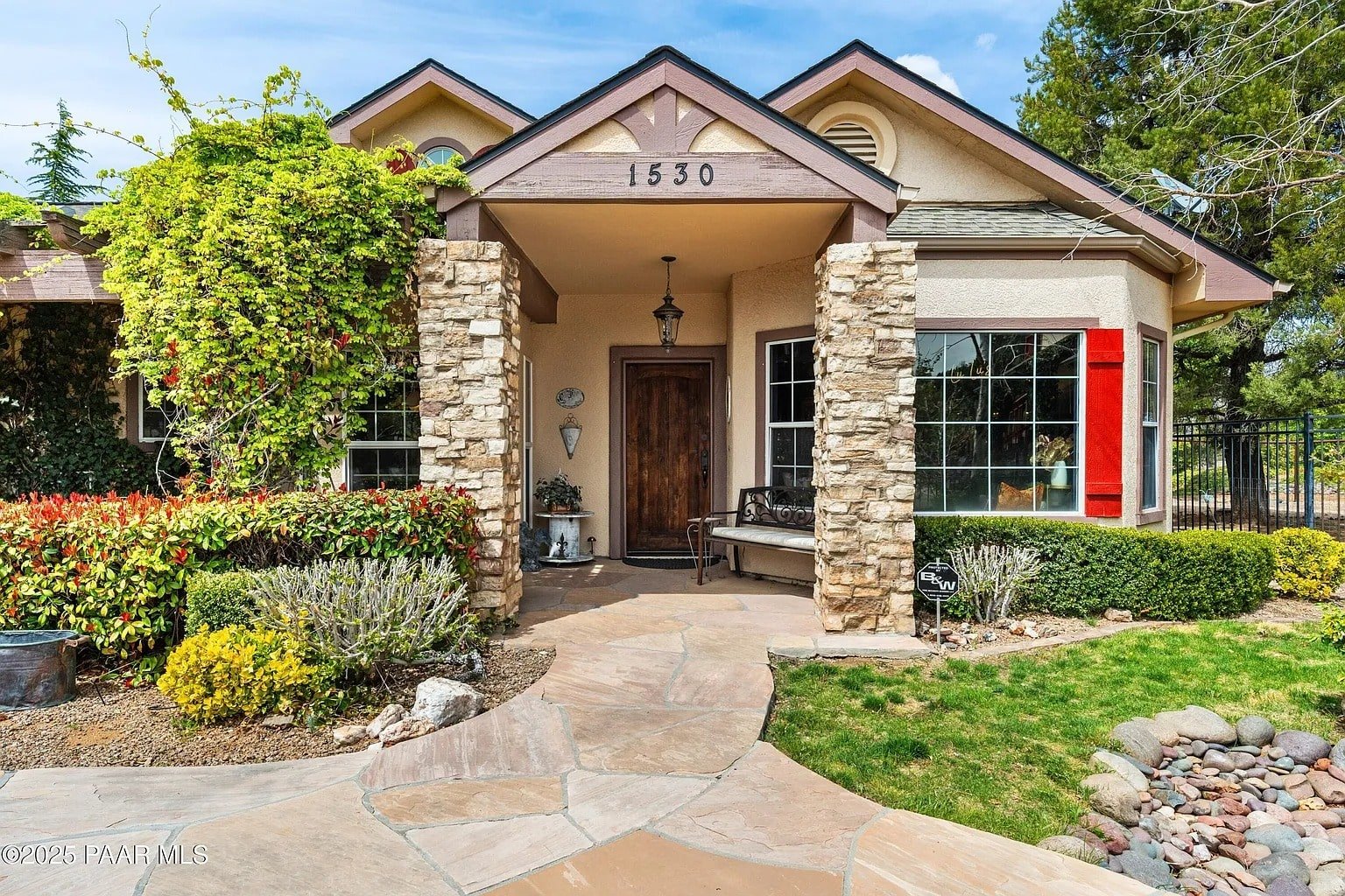 Single-story house with stone accents, red shutters, and a wooden door, surrounded by greenery.