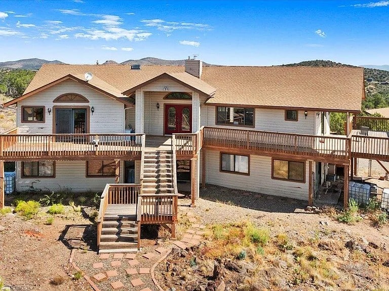 Two-story house with wooden deck, stairs, and surrounding dry landscape under a clear blue sky.