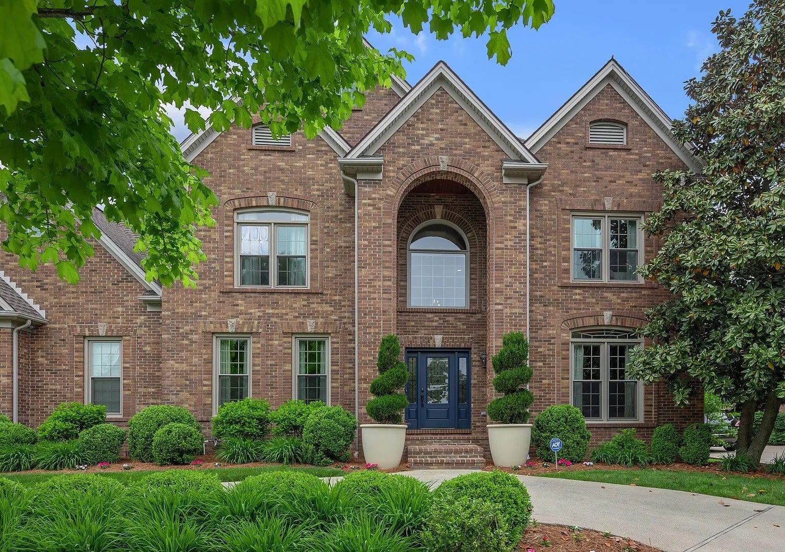 Large brick house with arched entrance, blue door, manicured shrubs, and surrounding greenery.