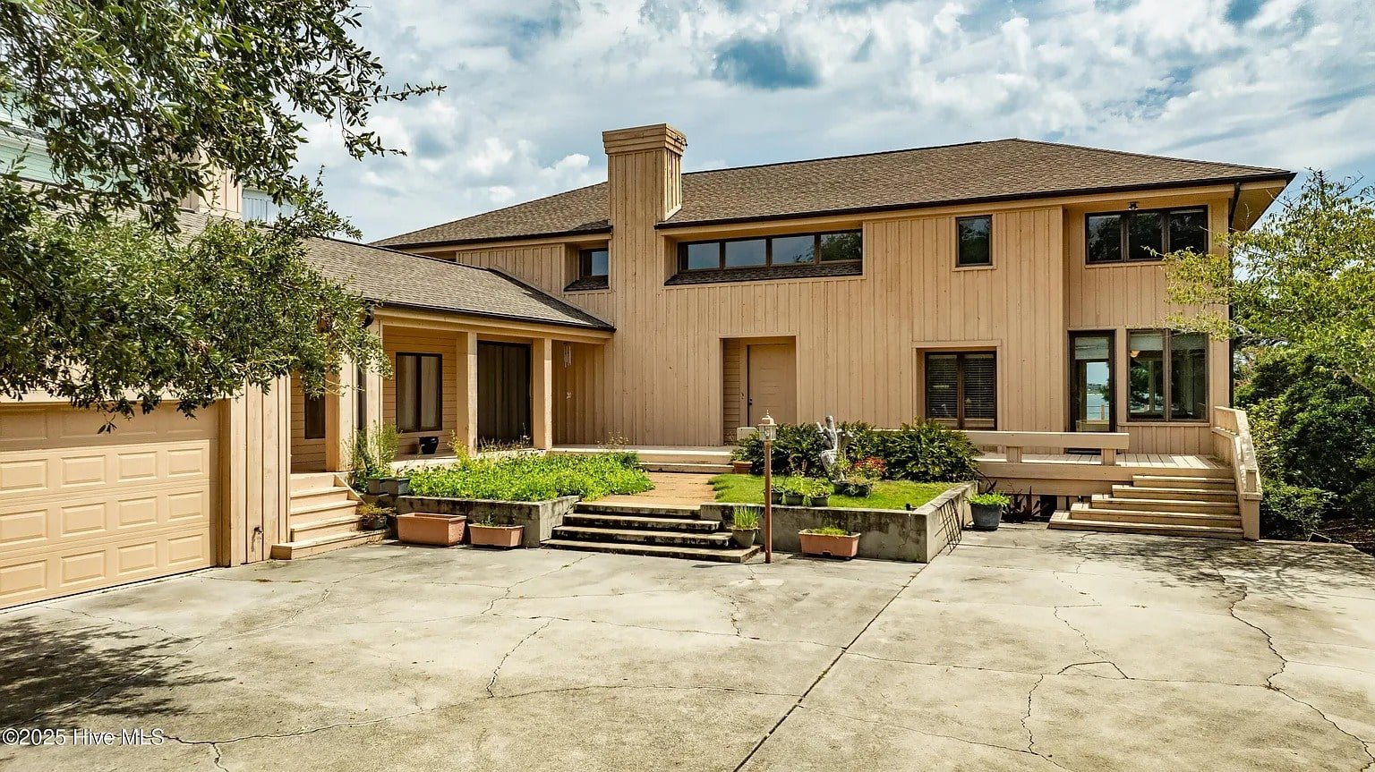 Two-story beige house with a large driveway, garage, and surrounding greenery under a partly cloudy sky.