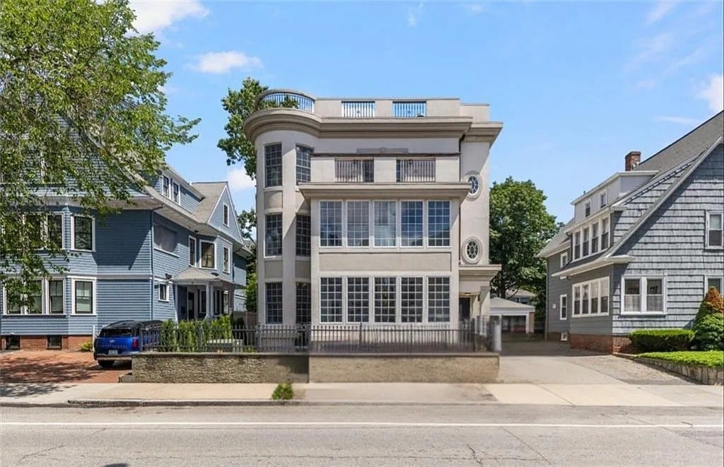 Modern three-story house with large windows, flanked by traditional homes, on a suburban street.