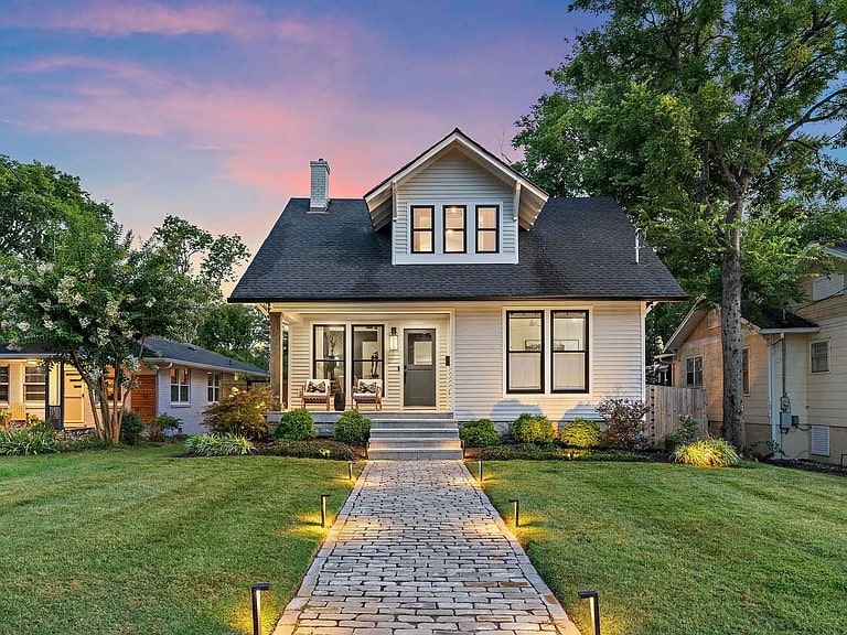 Two-story house with a lit pathway, surrounded by trees and a well-kept lawn at sunset.