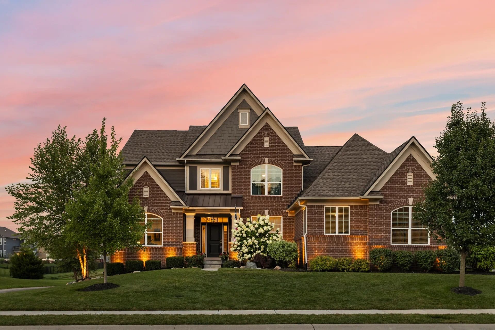 Brick house with peaked roofs, lit exterior, and a well-maintained lawn at sunset.