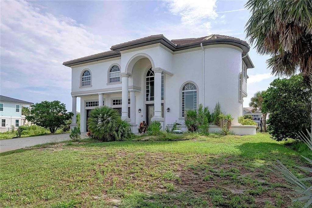 Large two-story white house with arched entrance, surrounded by greenery and palm trees.