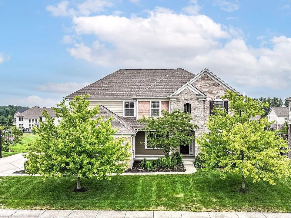 Two-story suburban house with a well-maintained lawn and trees, under a partly cloudy sky.