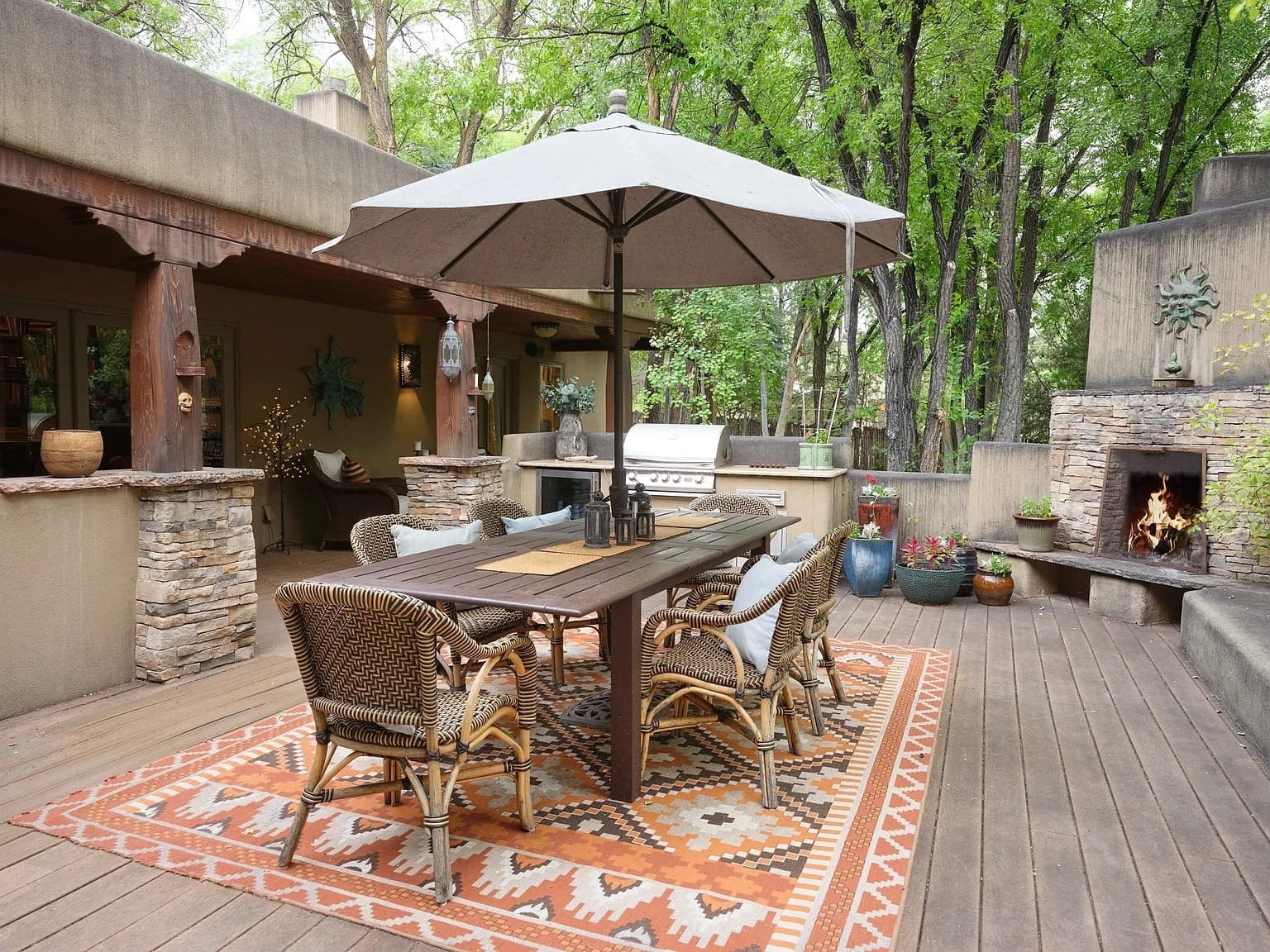 Outdoor patio with dining table, chairs, large umbrella, fireplace, and surrounding greenery.
