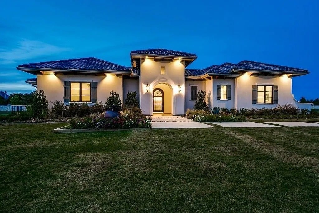 Modern single-story house with illuminated entrance, surrounded by a well-maintained lawn at dusk.