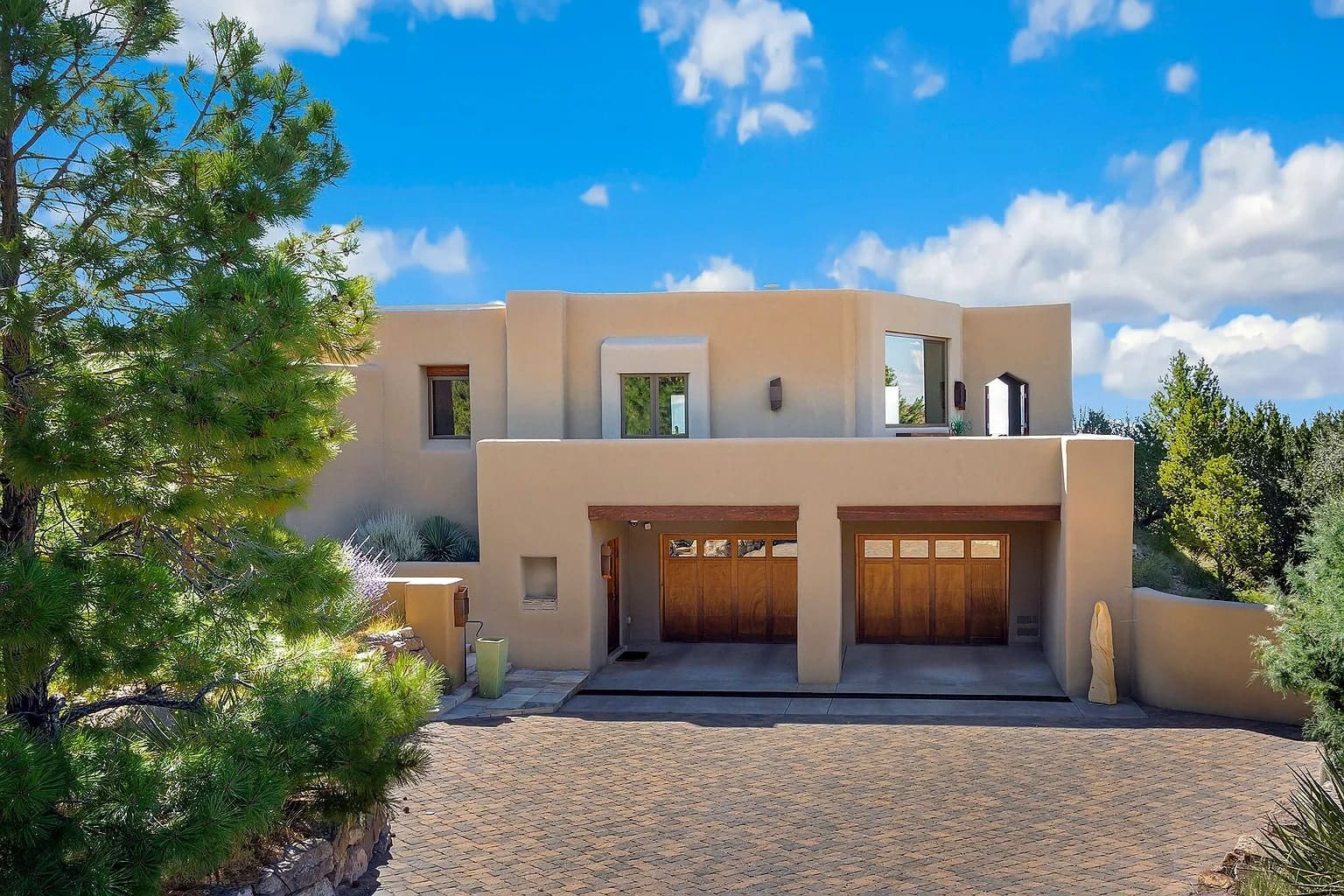 Modern adobe-style house with large wooden garage doors, surrounded by trees under a blue sky.