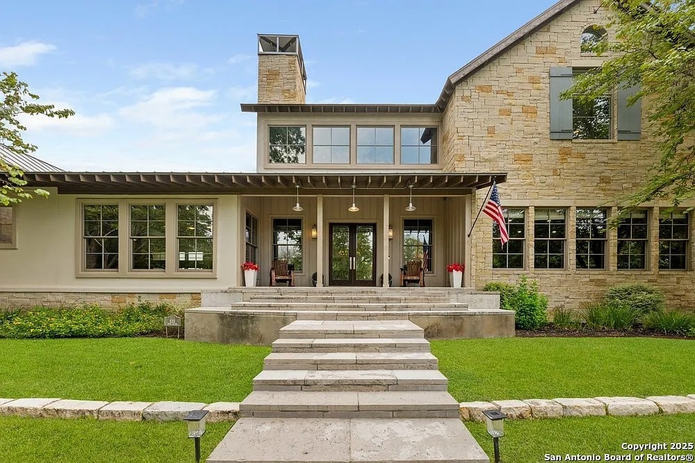 Modern stone house with large windows, front porch, and American flag. Green lawn and steps lead up.